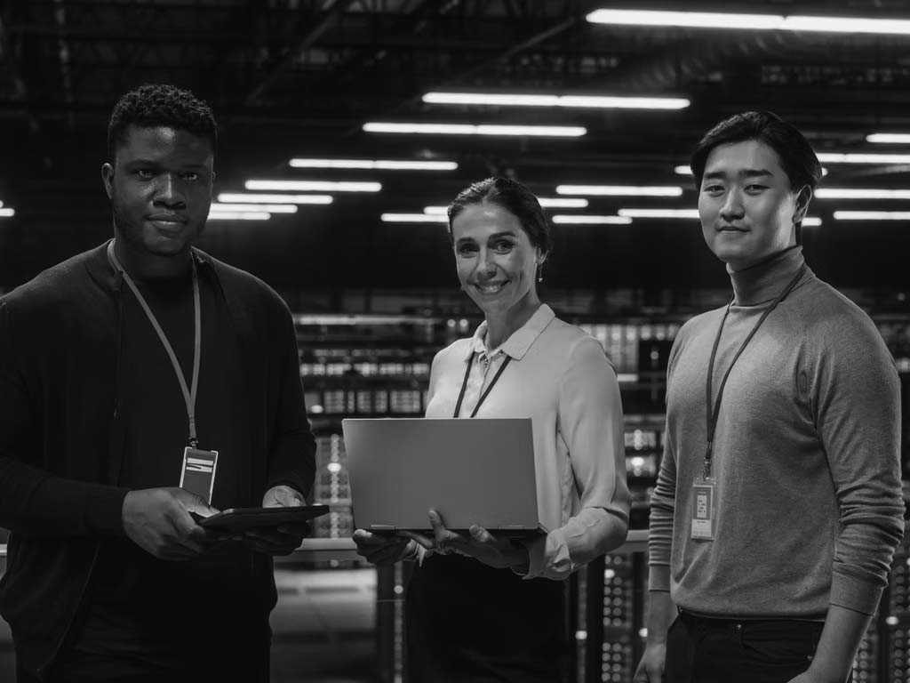 Three people stand in a high-tech server room, each holding a laptop or tablet. Wearing ID badges and business-casual attire, they smile confidently, embodying cyber security as blue server lights glow in the background.