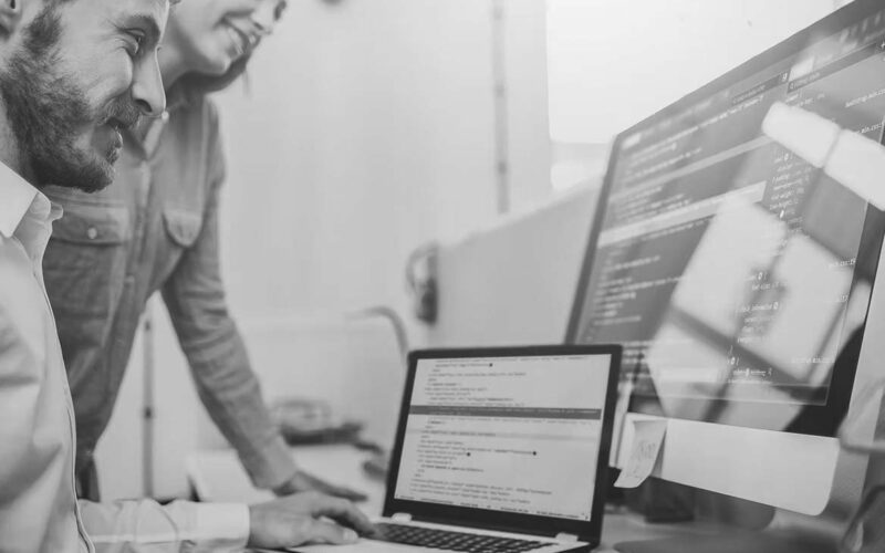 Two people are smiling while looking at computer screens displaying code. One person types on a laptop, while the other stands beside them, discussing tech solutions and ensuring compliance with guidelines on the desktop monitor.