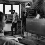 Several people are working in a modern office with exposed brick walls. One person types at a computer in the foreground, while three others discuss seamless transitions for deployment solutions on a large screen in the background.