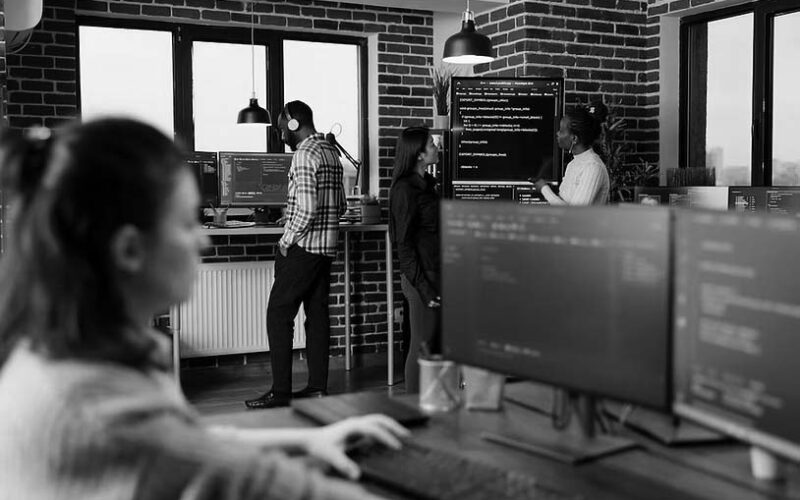 Several people are working in a modern office with exposed brick walls. One person types at a computer in the foreground, while three others discuss seamless transitions for deployment solutions on a large screen in the background.