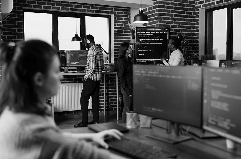 Several people are working in a modern office with exposed brick walls. One person types at a computer in the foreground, while three others discuss seamless transitions for deployment solutions on a large screen in the background.