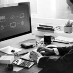 A person in a suit sits at a desk using a computer displaying a network diagram, researching Migration & Deployment Solutions from Pavelcomm. The desk has books, a calculator, wallet, smartphone, coffee, and plant. Large windows are in the background—Portland skyline visible.