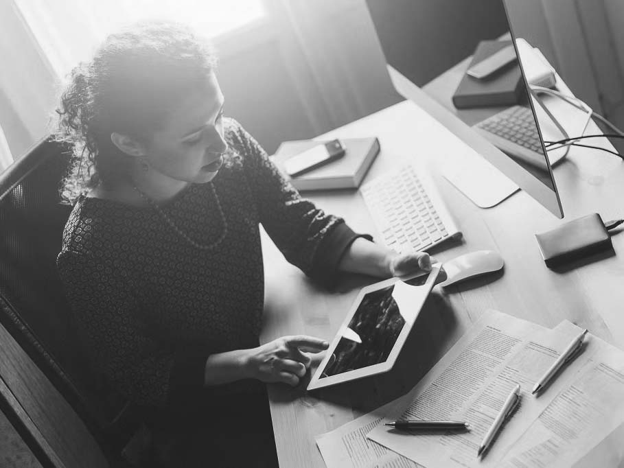 A woman sits at a desk using a tablet, with a desktop computer, keyboard, mouse, documents, and books around her—part of the growing at-home workforce—while sunlight streams through a window, illuminating her efficient workspace.