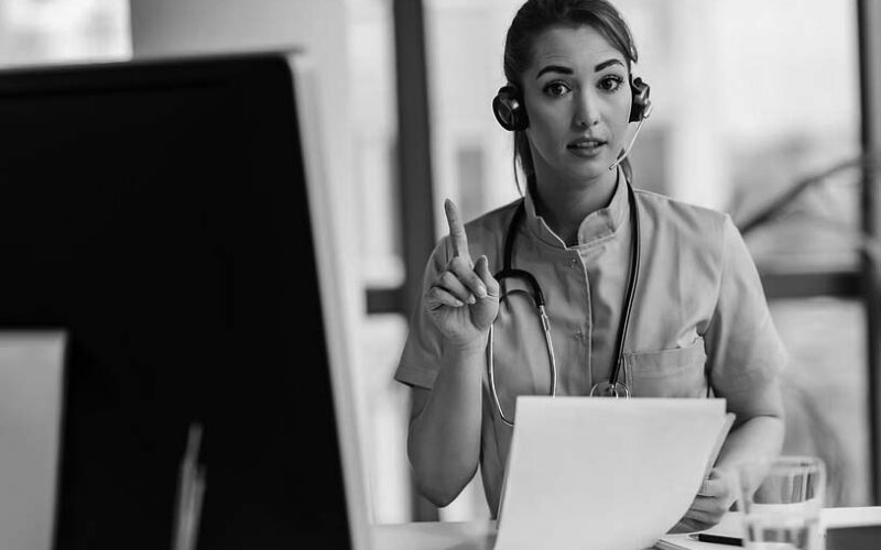 A female healthcare professional in blue scrubs and a headset holds up one finger while looking at a computer screen, suggesting she is optimizing patient care through expert advice during a virtual consultation.
