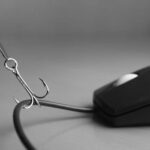 A fishing hook is caught on the cord of a computer mouse, symbolizing phishing or internet scams, with a focus on Phishing Prevention against a blurred background.