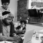 A man works on a laptop at a kitchen table—balancing business growth and IT consulting services—while a young boy sits on his shoulders and another child pulls his arm, creating a busy, multitasking scene in a home setting.