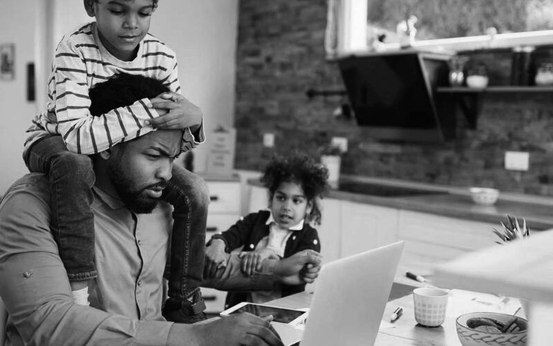 A man works on a laptop at a kitchen table—balancing business growth and IT consulting services—while a young boy sits on his shoulders and another child pulls his arm, creating a busy, multitasking scene in a home setting.