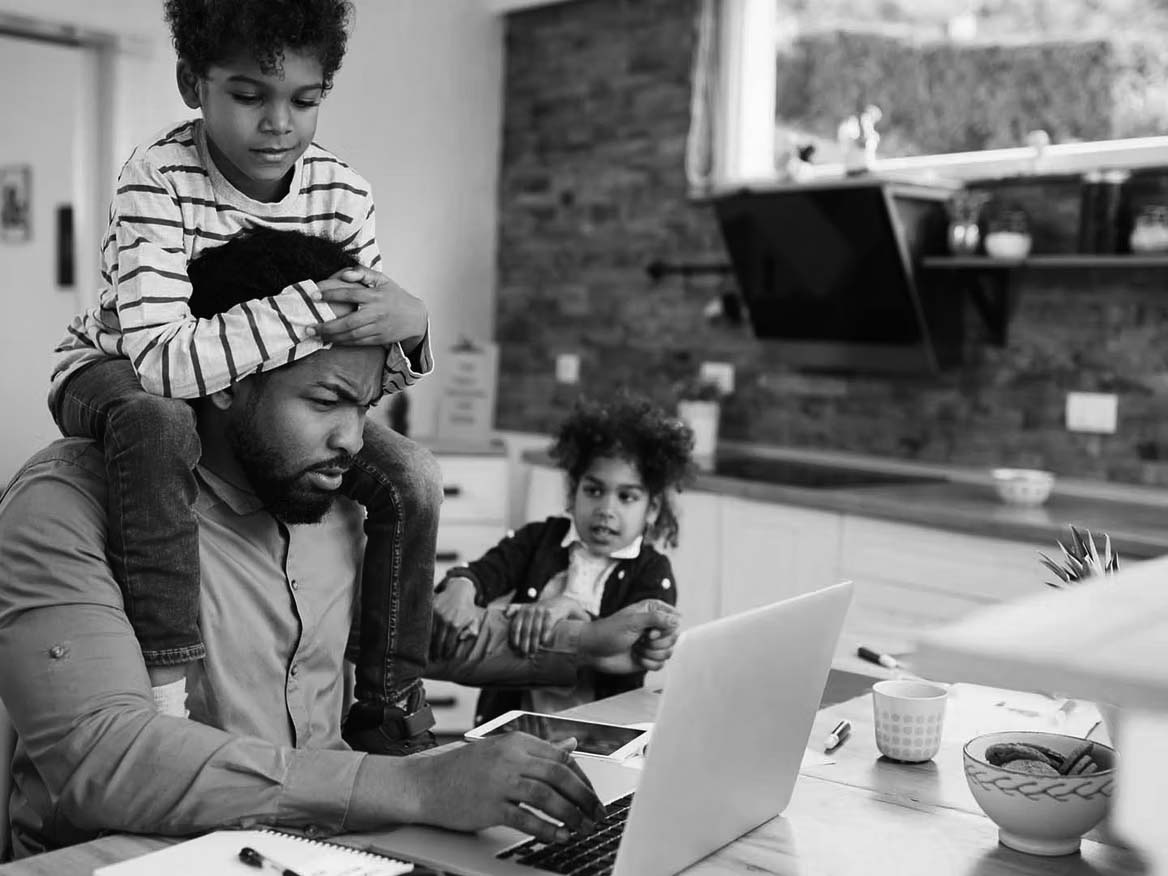 A man works on a laptop at a kitchen table—balancing business growth and IT consulting services—while a young boy sits on his shoulders and another child pulls his arm, creating a busy, multitasking scene in a home setting.