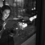 A woman stands by a window at night, using productivity tools on her digital tablet. City lights and traffic beyond create colorful reflections, capturing the essence of the modern workforce in motion.