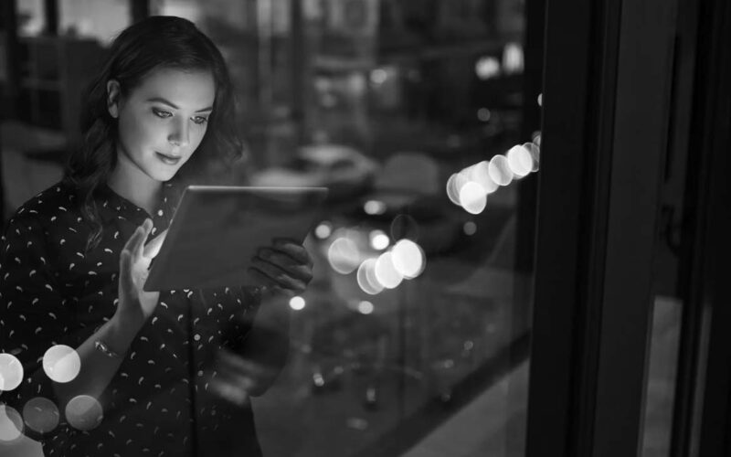 A woman stands by a window at night, using productivity tools on her digital tablet. City lights and traffic beyond create colorful reflections, capturing the essence of the modern workforce in motion.
