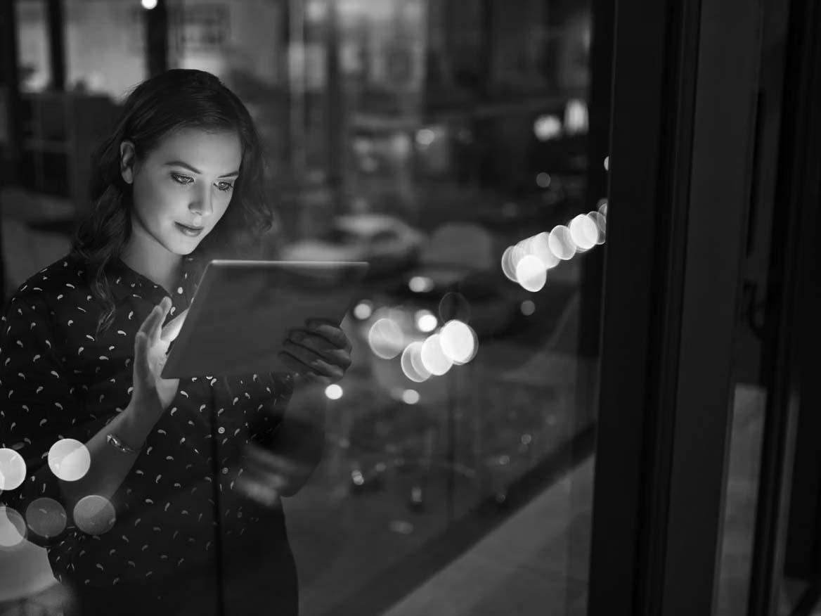 A woman stands by a window at night, using productivity tools on her digital tablet. City lights and traffic beyond create colorful reflections, capturing the essence of the modern workforce in motion.