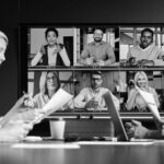 Four people sit at a conference table with documents and laptops, using fresh technology tools to participate in a video call with six colleagues displayed on a large screen in a virtual meeting.