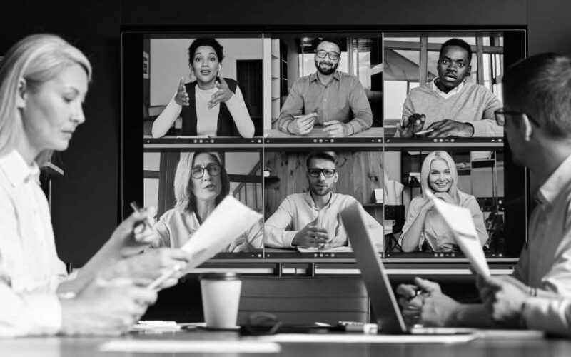 Four people sit at a conference table with documents and laptops, using fresh technology tools to participate in a video call with six colleagues displayed on a large screen in a virtual meeting.