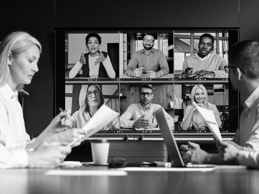 Four people sit at a conference table with documents and laptops, using fresh technology tools to participate in a video call with six colleagues displayed on a large screen in a virtual meeting.