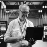 A man wearing glasses and a white shirt stands in a server room, using a laptop. As he monitors the illuminated servers, he provides expert virtualization services, streamlining operations for enhanced efficiency.