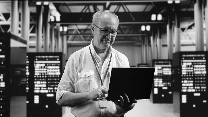 A man wearing glasses and a white shirt stands in a server room, using a laptop. As he monitors the illuminated servers, he provides expert virtualization services, streamlining operations for enhanced efficiency.