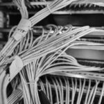 Close-up of numerous white low voltage cabling organized and bundled, connected to a server rack, with a blue-purple lighting effect illuminating the wires and equipment.