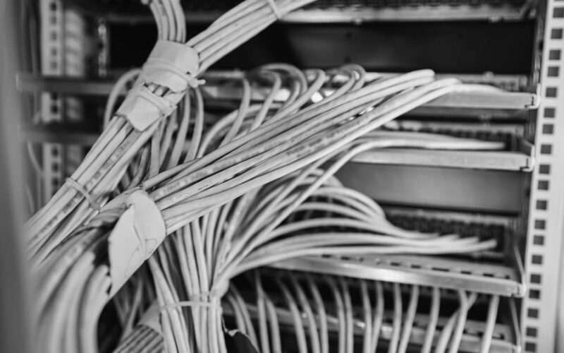 Close-up of numerous white low voltage cabling organized and bundled, connected to a server rack, with a blue-purple lighting effect illuminating the wires and equipment.