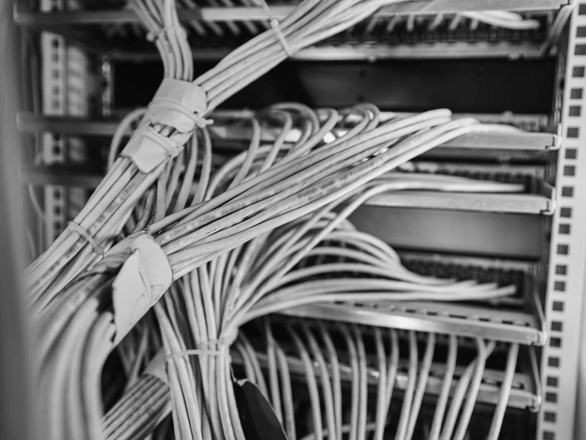 Close-up of numerous white low voltage cabling organized and bundled, connected to a server rack, with a blue-purple lighting effect illuminating the wires and equipment.