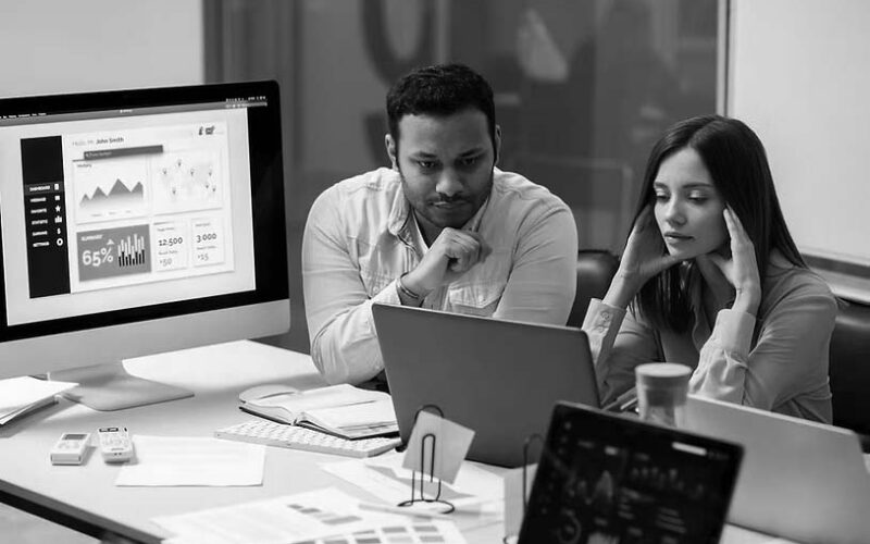 Two people sit at a desk looking intently at a laptop screen, surrounded by papers and digital devices displaying graphs and charts, suggesting a collaborative analysis session on IT support costs for small businesses in an office setting.