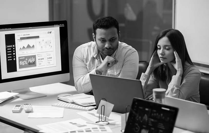 Two people sit at a desk looking intently at a laptop screen, surrounded by papers and digital devices displaying graphs and charts, suggesting a collaborative analysis session on IT support costs for small businesses in an office setting.