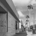 A row of storefronts along a sidewalk, some windows and doors boarded up with plywood, illustrates natural disaster preparedness. Palm trees and streetlights line the street under a partly cloudy sky.