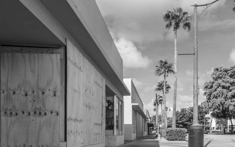 A row of storefronts along a sidewalk, some windows and doors boarded up with plywood, illustrates natural disaster preparedness. Palm trees and streetlights line the street under a partly cloudy sky.