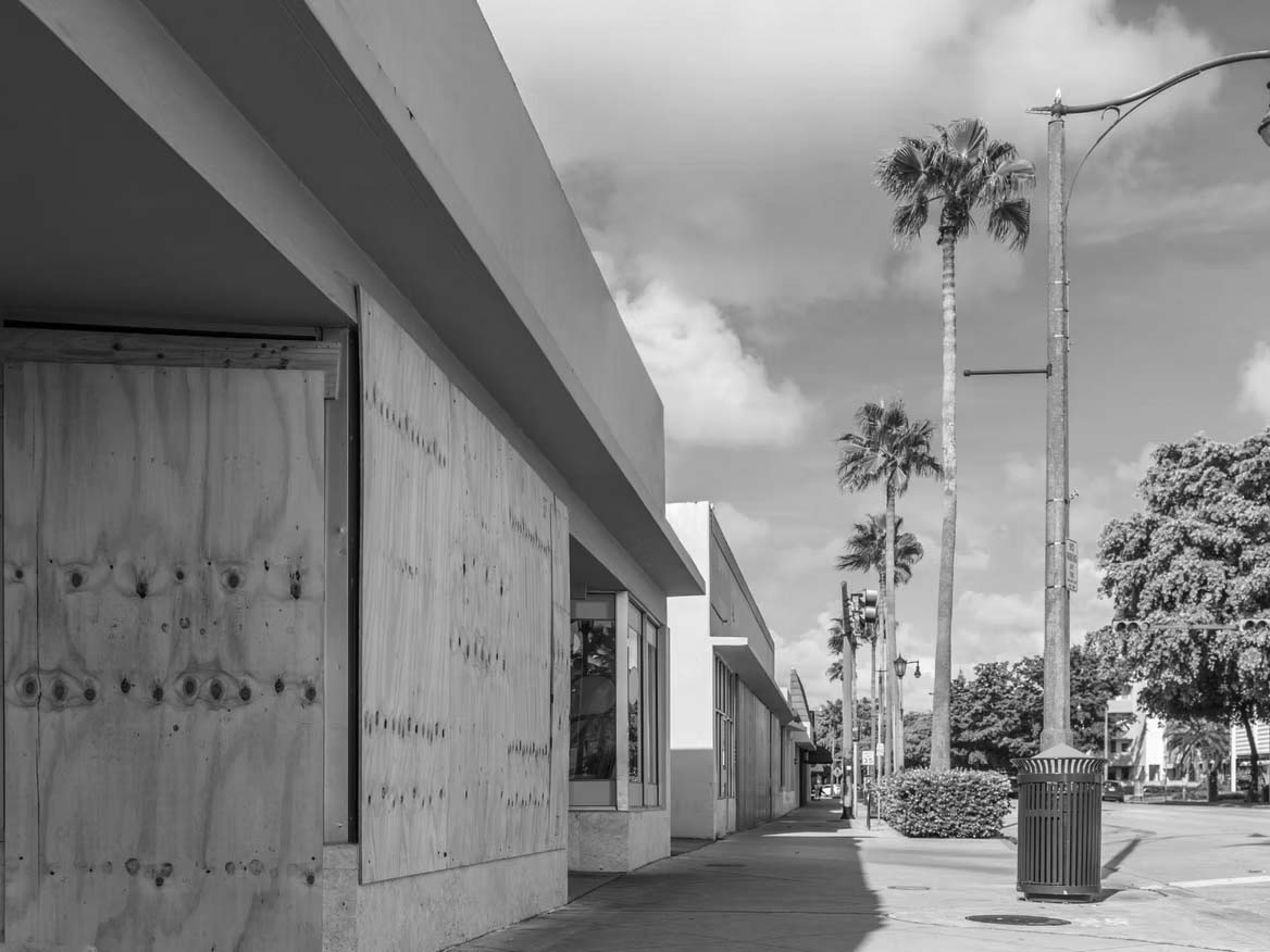 A row of storefronts along a sidewalk, some windows and doors boarded up with plywood, illustrates natural disaster preparedness. Palm trees and streetlights line the street under a partly cloudy sky.