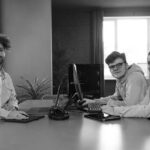 Three young adults sit at a desk with computers and headphones, looking at the camera in a bright room with large windows and green plants—showcasing how managed IT services are essential for your company’s modern workspace.