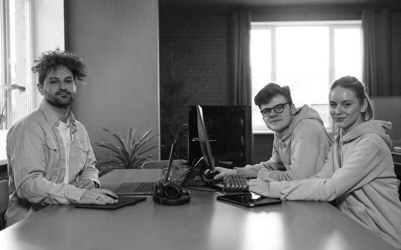 Three young adults sit at a desk with computers and headphones, looking at the camera in a bright room with large windows and green plants—showcasing how managed IT services are essential for your company’s modern workspace.