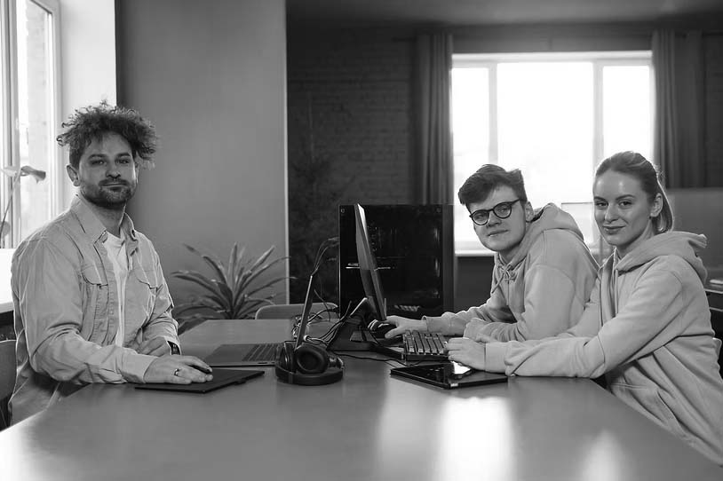 Three young adults sit at a desk with computers and headphones, looking at the camera in a bright room with large windows and green plants—showcasing how managed IT services are essential for your company’s modern workspace.