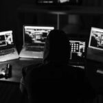 A person wearing a hood sits at a desk in Portland, surrounded by four laptops displaying code—suggesting hacking activity tied to the Dark Web. A handgun and smartphone rest on the table in the dimly lit room.
