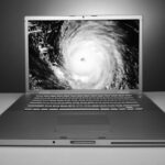 A silver laptop on a white surface displays a satellite image of a hurricane with a visible eye on its screen, highlighting the risk of data disaster for any small business, set against a blue gradient background.
