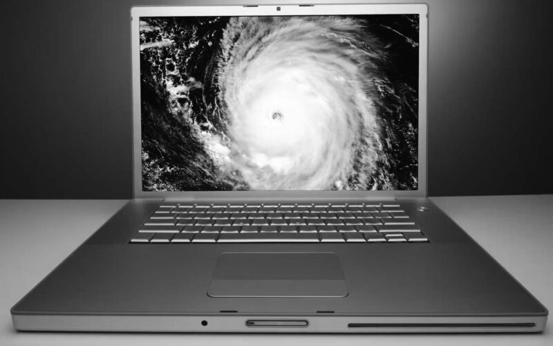 A silver laptop on a white surface displays a satellite image of a hurricane with a visible eye on its screen, highlighting the risk of data disaster for any small business, set against a blue gradient background.