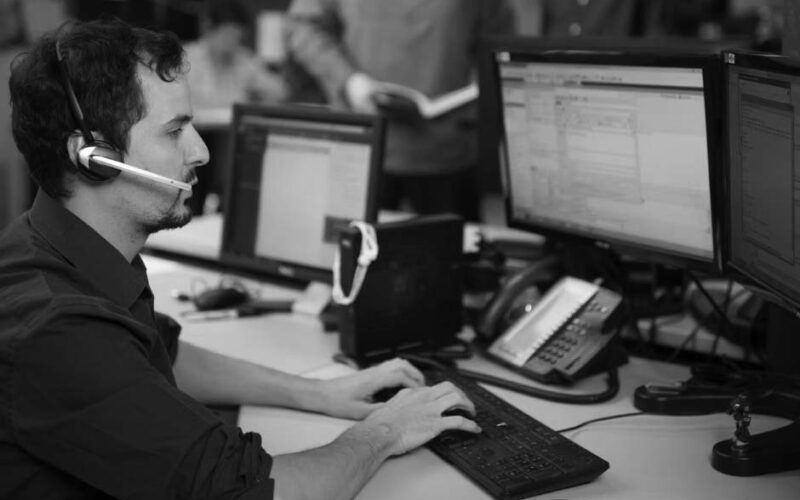 A man wearing a headset sits at a desk with three computer monitors, typing on a keyboard in an office. Other people are visible in the background, collaborating and talking, creating an optimized work environment.