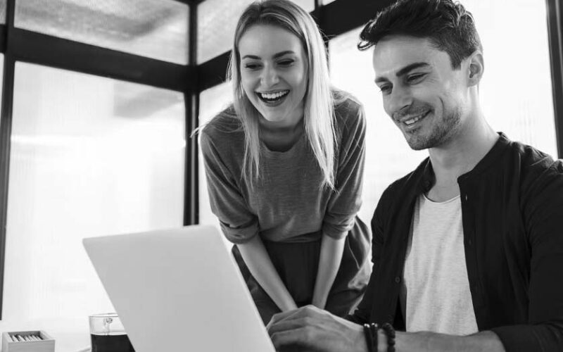A man and a woman smile as they look at a laptop screen together in a modern office with sleek modern cabling. The man is seated and typing, while the woman stands beside him, leaning forward and laughing.