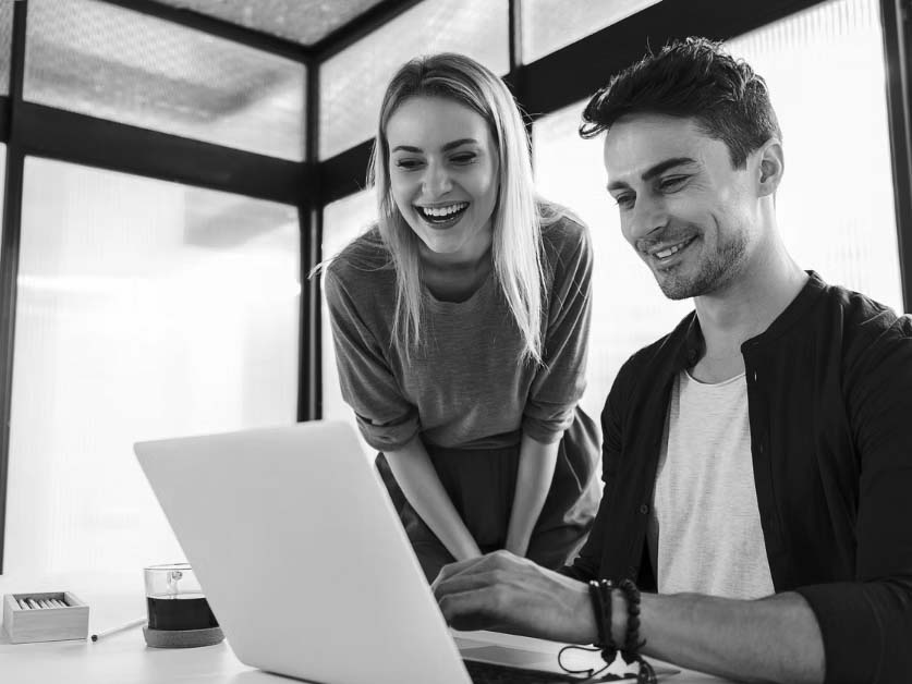 A man and a woman smile as they look at a laptop screen together in a modern office with sleek modern cabling. The man is seated and typing, while the woman stands beside him, leaning forward and laughing.