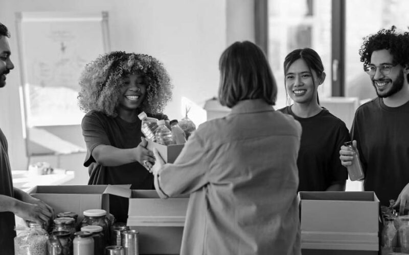 Four volunteers in green shirts pack food items into boxes, offering IT solutions for nonprofits, and hand them to a woman in a brown jacket at an indoor food bank. They are smiling and surrounded by jars, cans, and bottles on the table.