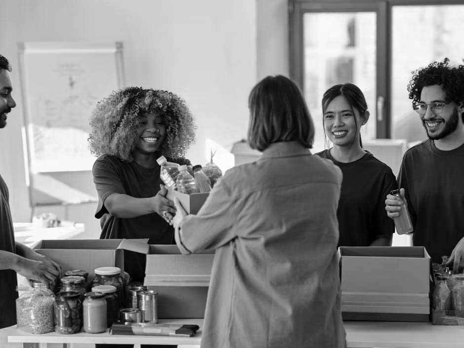Four volunteers in green shirts pack food items into boxes, offering IT solutions for nonprofits, and hand them to a woman in a brown jacket at an indoor food bank. They are smiling and surrounded by jars, cans, and bottles on the table.
