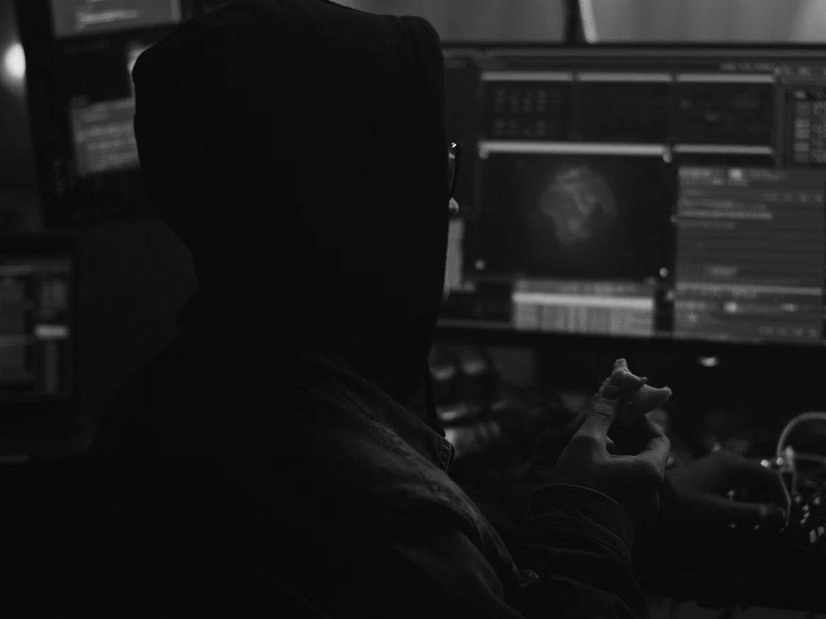 A person in a hoodie sits in front of multiple computer screens displaying code and a digital map with a target alert, holding a partially eaten sandwich in one hand. The room is dimly lit, giving a secretive atmosphere.