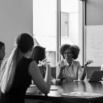 Five people sit around a conference table in discussion. One woman gestures while speaking about network security, while others listen attentively. In the background, a chart with graphs is displayed on a whiteboard near large windows.