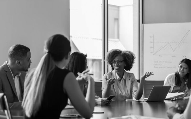 Five people sit around a conference table in discussion. One woman gestures while speaking about network security, while others listen attentively. In the background, a chart with graphs is displayed on a whiteboard near large windows.