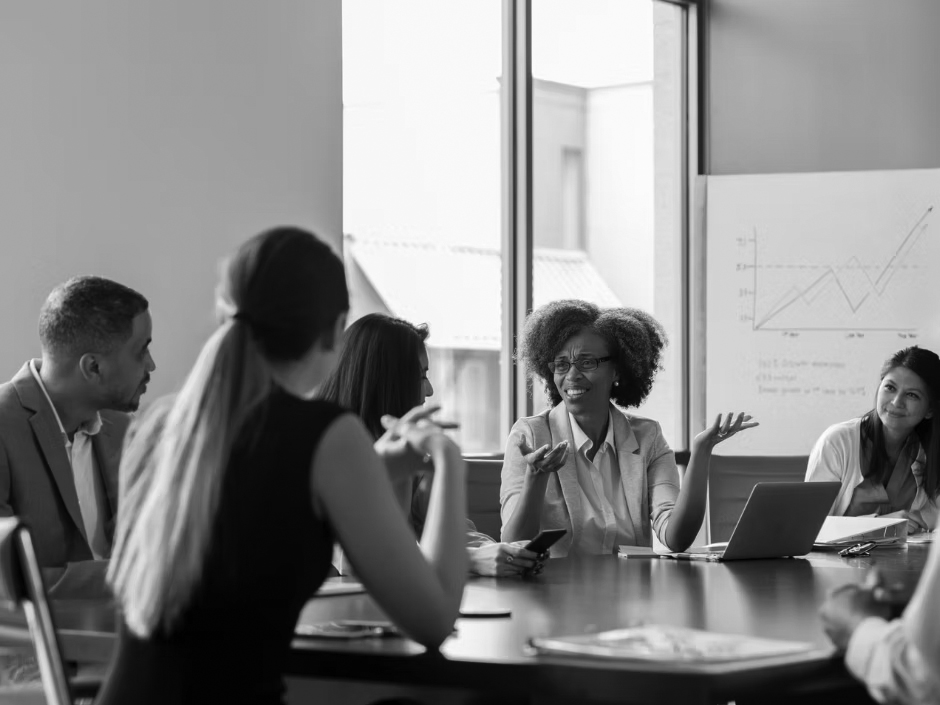 Five people sit around a conference table in discussion. One woman gestures while speaking about network security, while others listen attentively. In the background, a chart with graphs is displayed on a whiteboard near large windows.