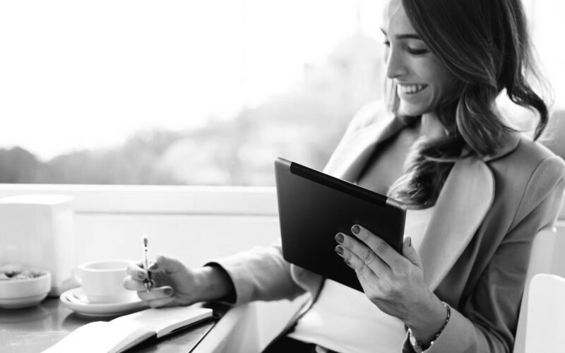 A woman sits at a table with a coffee cup, smiling while holding a tablet in one hand and writing notes for her company self-assessment in a notebook with the other. She wears a light blazer and has long hair.