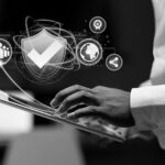 A person uses a tablet with a digital overlay of a shield checkmark icon and various technology and security symbols, illustrating IT security and measures to secure your IT against threats like ransomware.