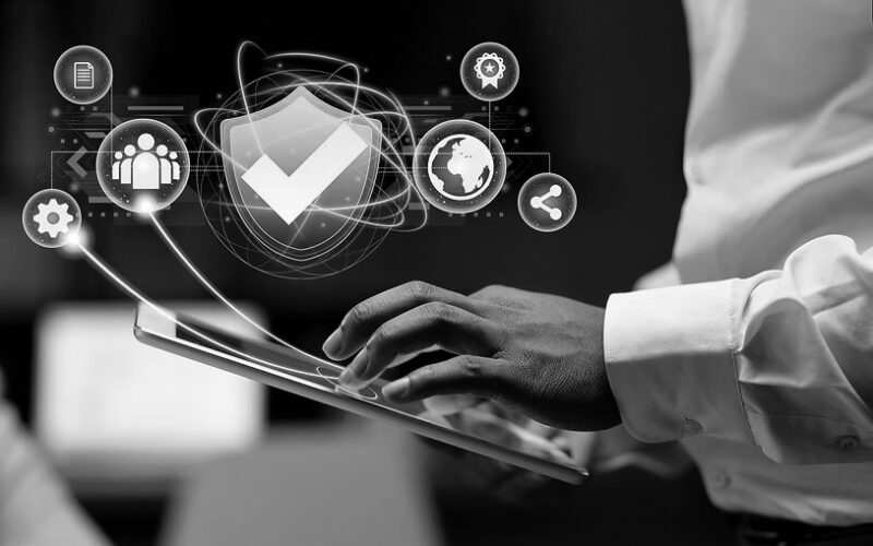 A person uses a tablet with a digital overlay of a shield checkmark icon and various technology and security symbols, illustrating IT security and measures to secure your IT against threats like ransomware.