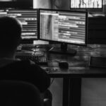 A person sits at a desk working on two computer monitors displaying lines of code related to cybersecurity in a dimly lit room. A keyboard, mouse, and tablet are also on the desk.