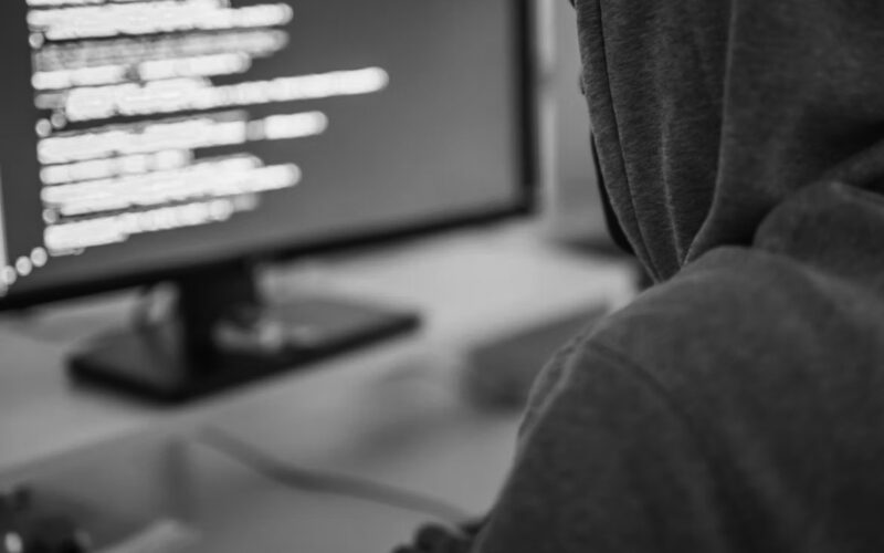 A person in a gray hoodie sits at a desk, facing a computer monitor displaying lines of code with a blue background. The scene suggests programming or cybersecurity activities—don’t wait to protect yourself against online threats.