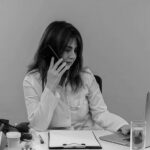 A female doctor in a white coat talks on the phone with the Emergency Help Desk while working on a laptop at a desk with medical tools, papers, and a glass of water, set against an orange background.