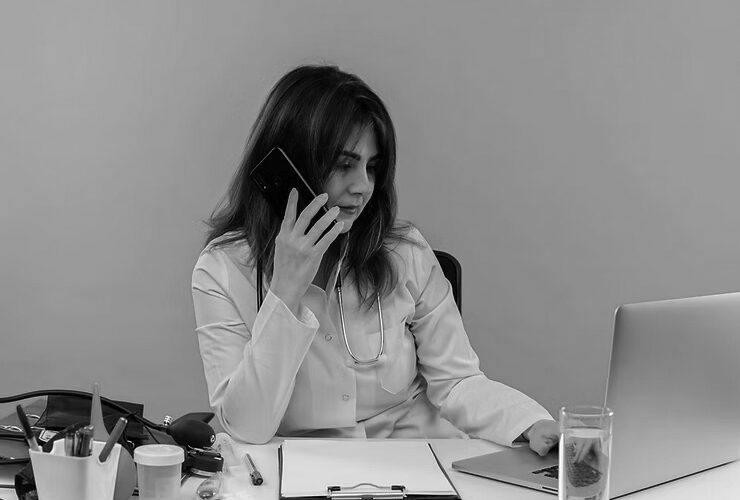 A female doctor in a white coat talks on the phone with the Emergency Help Desk while working on a laptop at a desk with medical tools, papers, and a glass of water, set against an orange background.